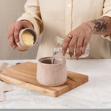 Person pouring Water from a glass container into a ceramic mug on a wooden cutting board.