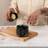 Person pouring a water from a bottle into a black marble cup on a wooden cutting board.