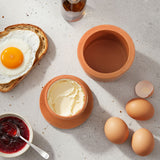 Breakfast scene with eggs, toast, and a terracotta container on a light surface.