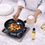 A chef in a white uniform adds salt from a marble container to vegetables cooking in a black non-stick frying pan on a gas stove, set on a white marble countertop.