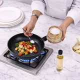 A person in a white chef coat adds salt from a marble salt container to vegetables cooking in a black frying pan on a gas stove, set on a white marble countertop with a bottle of oil nearby.