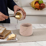 Person spreading butter on bread using a ceramic container and knife, with a kitchen setting in the background.