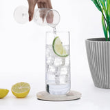 Person pouring a clear liquid into a glass with ice and a lime slice, with a potted plant in the background.