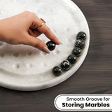 A hand with a gold ring places a smooth black marble onto a white marble tray with grooves, with more black marbles lined up on the side. A text overlay at the bottom reads 'Smooth Groove for Storing Marbles'.