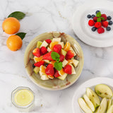 Fruit salad in a bowl on a marble surface with additional fruits and a glass of lemonade.