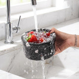 Collapsible colander with strawberries being rinsed under running water in a kitchen sink.