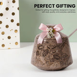 A brown marbled jar with a lid, decorated with a pink satin bow and small white flowers, shown on a white surface with a mint green background and a white gift bag with gold dots in the side. Text at the top reads 'PERFECT GIFTING'.