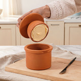 Person pouring a creamy substance from one terracotta container into another on a wooden cutting board.