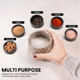 A hand holds a round stone salt and pepper grinder in the center, surrounded by small black bowls labeled Sugar, Pepper, Spices, Pink Salt, and Almonds on a light surface. Text at the bottom reads 'MULTI PURPOSE' with additional descriptive copy.