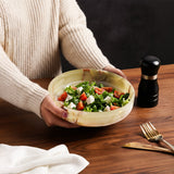 Person holding a bowl of salad on a wooden table with a pepper grinder and cutlery.