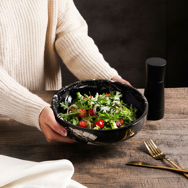 Person holding a black bowl of salad on a wooden table with a pepper grinder and cutlery.