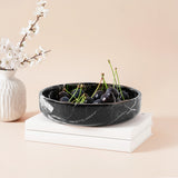 Black marble bowl with cherries on a beige surface with a white vase and branches.