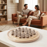 A round stone game board with multiple polished oceanic stones balls  arranged on it, placed on a light table in the foreground. The background shows a softly blurred living room with two people sitting on an orange-brown sofa, creating a lifestyle context.
