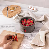 Person cutting strawberries on a wooden cutting board with a bowl of strawberries and eggs in the background.