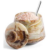 A decorative salt cellar made of marbled onyx  shown open with coarse pink salt inside and a metal spoon resting in it. The matching marbled lid is placed in front on a clean white background.