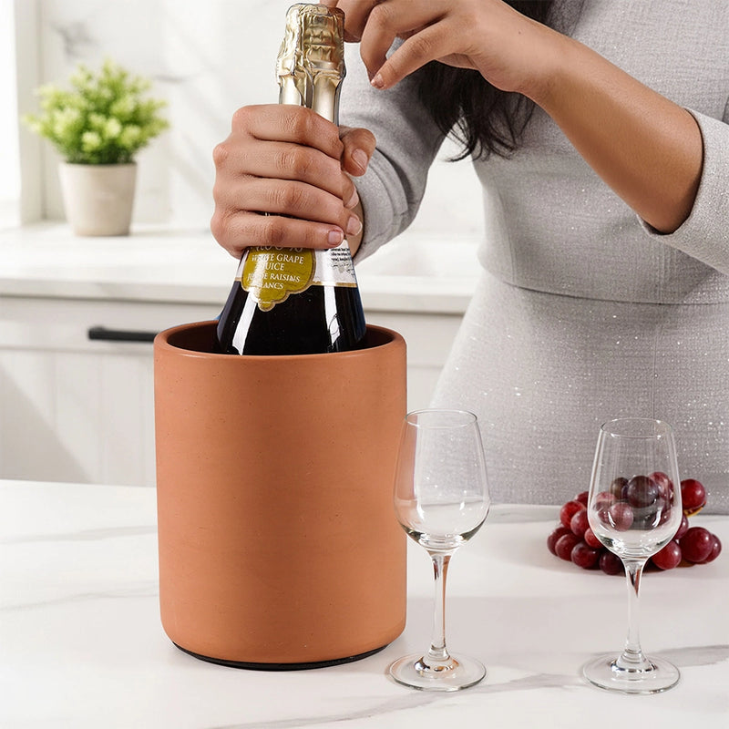 Person opening a bottle of champagne in a terracotta wine chiller with glasses and grapes on a table.