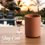 Person pouring red wine into a glass with a terracotta-colored mug on a wooden table outdoors.