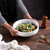 Person holding a white marble bowl filled with a green salad on a wooden table.