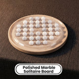 A polished marble solitaire board with white marble balls arranged on it, shown on a dark textured fabric background with a text label at the bottom that reads 'Polished Marble Solitaire Board'.