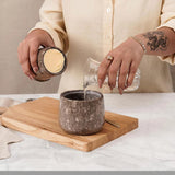 Person pouring a water from a jar into a stone cup on a wooden board.