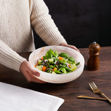 Person holding a bowl of salad on a wooden table with a pepper grinder and cutlery.