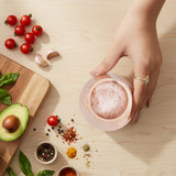 Hand holding a salt cellar with a wooden cutting board, tomatoes, garlic, and spices on a light wood surface.