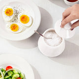 A white marble salt container with a matching lid is shown on a light table surface, with a hand lifting the lid. A spoon sits inside the container filled with white salt.