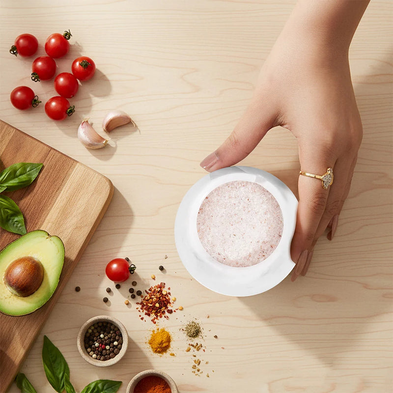 A hand wearing a gold ring holds a small white salt cellar  filled with pink Himalayan salt on a light wood table, surrounded by fresh ingredients like cherry tomatoes, garlic, avocado, basil, and spices.