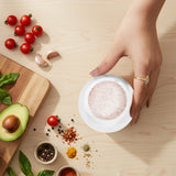 A hand wearing a gold ring holds a small white salt cellar  filled with pink Himalayan salt on a light wood table, surrounded by fresh ingredients like cherry tomatoes, garlic, avocado, basil, and spices.