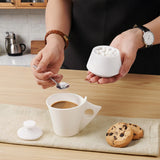 A person holds a small white salt cellar and a spoon over a white ceramic coffee mug on a wooden table, with chocolate chip cookies nearby.