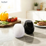 Two marble-style salt and pepper shakers, one white and one black, on a light gray marble countertop in a bright kitchen setting with food props in the background.