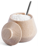 A beige stone salt and pepper jar with a matching lid leaning in front, filled with coarse white salt and topped with a metal spoon, on a white background.