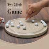 Two hands playing with a traditional game on a wooden table, featuring the text 'Two Minds One Timeless Game'.
