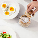 A hand is sprinkling coarse salt from a small round  container onto a spoonful, set on a white tablecloth next to plates of food including hard-boiled eggs and a salad.