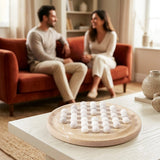 A round marble board game with white marble-like pieces arranged on a light wood table in a bright living room. Two people are seated on a rust-orange sofa in the background, softly blurred.