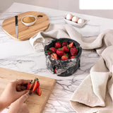 A round black marble strawberry cake stand filled with fresh strawberries sits on a white marble countertop. A person's hands are visible slicing strawberries on a wooden cutting board in the foreground, with a beige cloth draped around the stand and eggs in a white dish in the background.