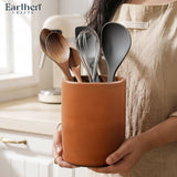 Person holding a terracotta utensil holder with kitchen utensils, branded 'Earthen Crafts'.