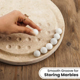 A hand placing a smooth white marble balls onto a round travertine board  arranged along the edge, on a wooden surface. Text overlay reads 'Smooth Groove for Storing Marbles'.