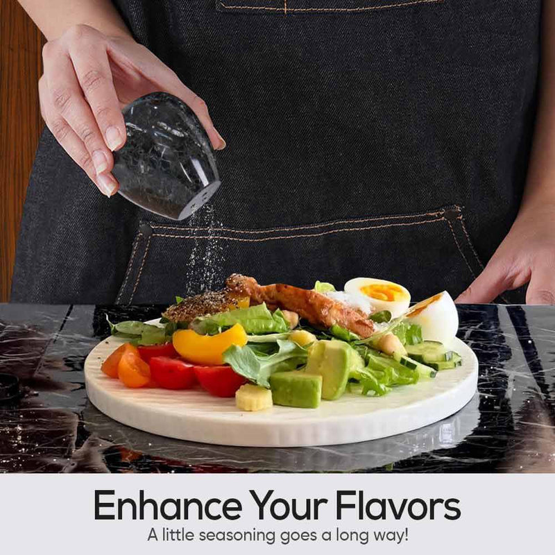 Person seasoning a salad with a black salt grinder on a marble cutting board.