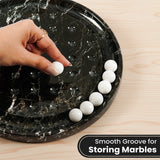 A hand places a white marble ball into a black marble board , with several white marbles lined up on the side. The image is set on a light wood background with a text overlay that reads 'Smooth Groove for Storing Marbles'.