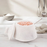 A white marble-style salt container with its lid off, filled with pink Himalayan salt and a metal spoon, set on a light marble countertop with a neutral kitchen background.