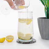 Pouring water into a glass with lemon slices on a white surface with a plant in the background.