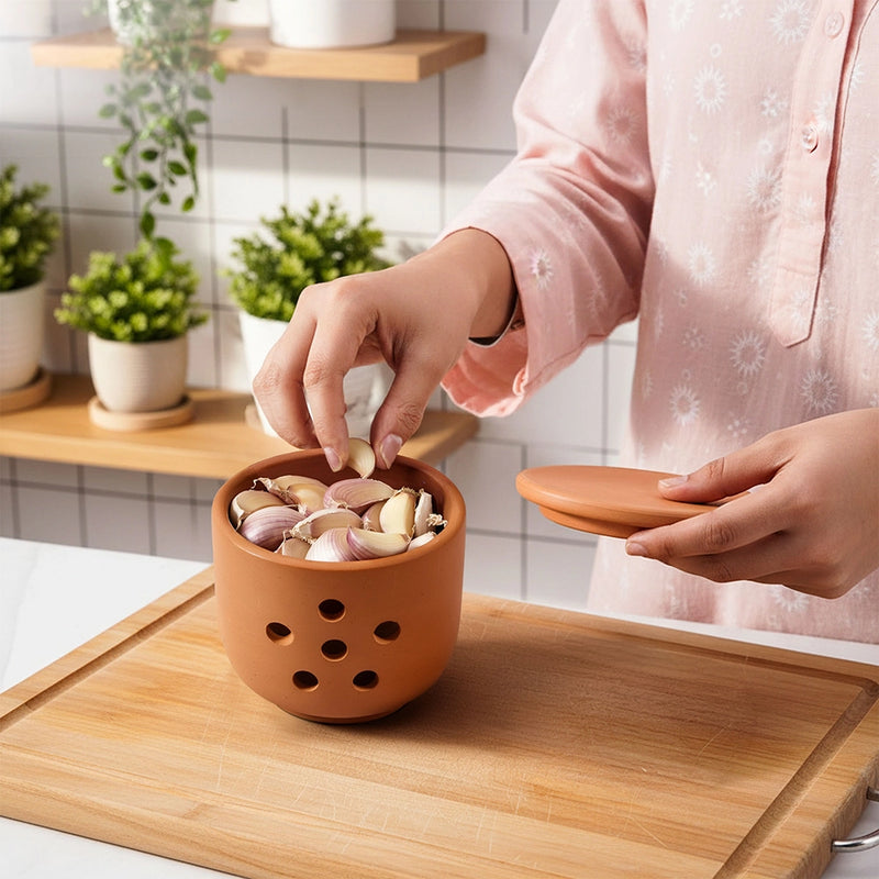 Person adding garlic cloves into a terracotta-colored garlic peeler on a wooden cutting board.