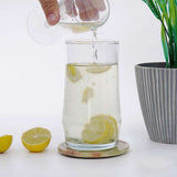 Pouring lemon slices into a glass of water on a green coaster plate placed on white surface with a plant in the background.