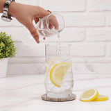 Pouring lemon slices into a glass of water on wood coaster plate placed on white surface with a plant in the background.