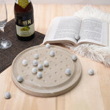 Round stone solitaire board with marble-like stones on a wooden surface with a bottle and book in the background.