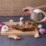 Person seasoning a roasted meat dish with salt on a wooden board, surrounded by garlic and onions.