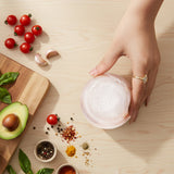 Hand holding a small container on a wooden surface with ingredients like tomatoes, garlic, and avocados.