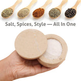 Hand holding a wooden container with salt, surrounded by wooden spoons with various spices on a white background.