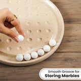 A hand placing a white marble balls into a beige stone board with circular grooves, with several white marbles lined up along the bottom edge on a wooden surface. Text in the corner reads 'Smooth Groove for Storing Marbles.
